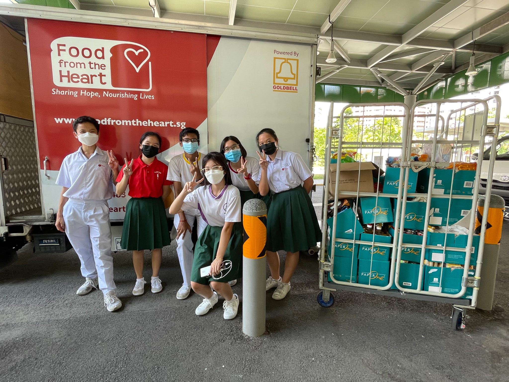 Six masked students in uniforms pose with peace signs near a Food from the Heart charity truck and a cart of donated boxes.